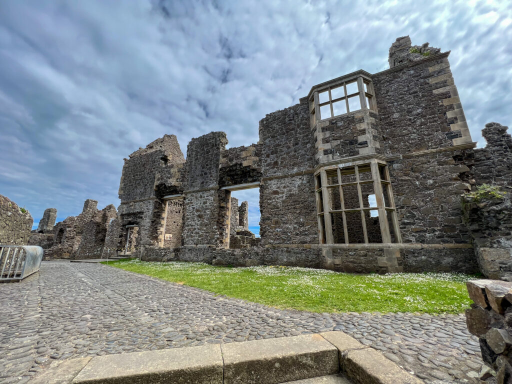 An interior shot of Dunluce Castle, said to be the inspiration for Prince Caspian in Cair Paravel. Stone walkways and a strip of grass lead to the ruins of the manor house, with some of the bay window stone intact. The ruins of other castle structures can be seen in the background.