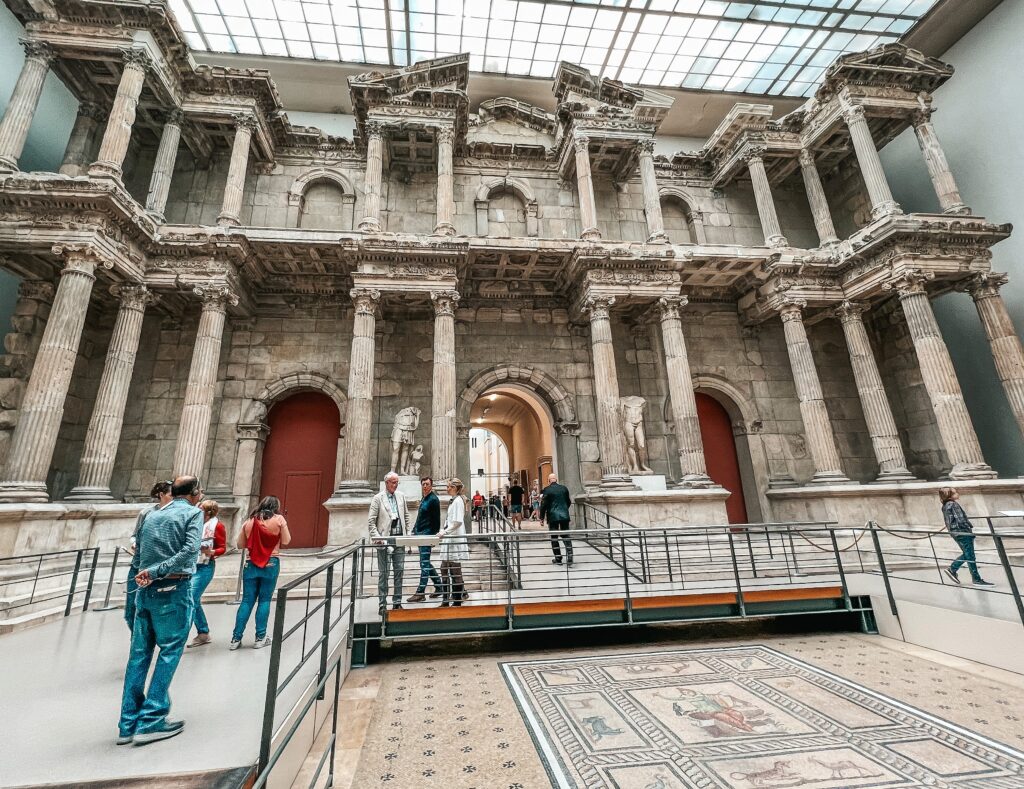 What is History? Inside the Pergamon Museum in Berlin, people look up at a two-story stone building facade structure with multiple classical columns.